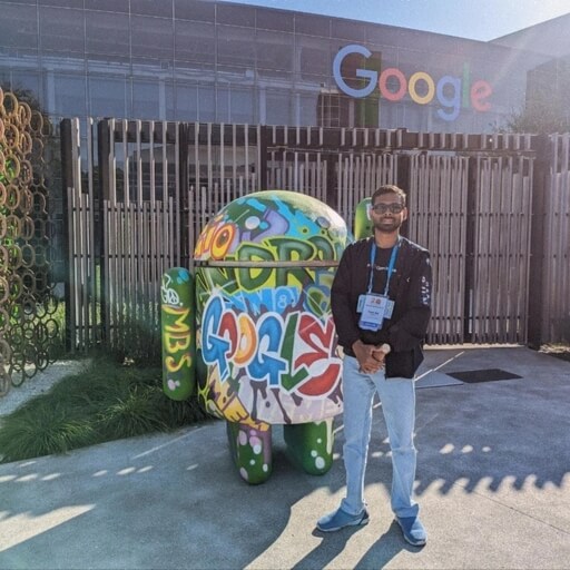 一名男子站在一尊色彩鲜艳、涂鸦着Android雕像旁边,背景是谷歌总部。男子戴着挂绳,天气晴朗。